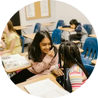 Clear communication Two young girls smiling and chatting at a classroom desk, practicing communication skills.